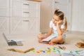 Tired little schoolgirl girl writing by looking to laptop screen during homeschooling sitting on floor at home. Royalty Free Stock Photo