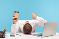 Tired man office worker lying face down on desk holding paper cup of coffee and showing thumbs up, need coffee to be ok, Royalty Free Stock Photo