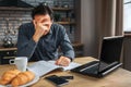 Tired man sit at table in kitchen. He cover face with hand. Man work. He has headache.