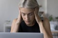 Tired mature blonde woman in casual touching head at laptop