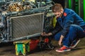 A tired mechanic in a blue protective suit is repairing a car radiator. Repair Service Concept.