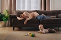 Tired mother sleeping on sofa while her toddler baby playing on floor