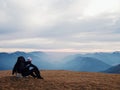 Tired out tourist in black with backpack is sitting on stone in meadow and watching into misty valley. Autumn in mountains.