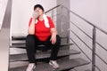 Tired overweight mature woman drinking water from bottle on stairs indoors