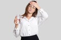 Tired overworked woman keeps hand on forehead, wears transparent glasses, black and white clothes, holds glasses of water, feels t