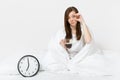 Tired sad woman sitting in bed with cup of coffee, round clock, white sheet, pillow, wrapping in blanket on white