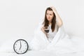 Tired sad woman sitting in bed with cup of coffee, round clock, white sheet, pillow, wrapping in blanket on white