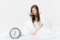 Tired sad woman sitting in bed with cup of coffee, round clock, white sheet, pillow, wrapping in blanket on white