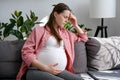 Tired sad young pregnant female suffering migraine sitting alone on sofa at home. Unhappy future mom holding painful head while Royalty Free Stock Photo
