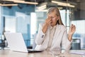 Tired senior woman dressed in office suit sitting at desk with laptop and touching forehead while having headache Royalty Free Stock Photo