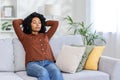 Tired and serious young African American woman sitting on sofa at home and resting, hands behind head and eyes closed