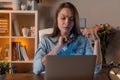 Tired serious young girl in jeans shirt working at home and use her laptop. The pen in the girl`s hands. The shelf with books is Royalty Free Stock Photo