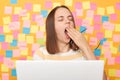 Tired sleepy bored young adult woman with brown hair working on laptop, keeps eyes closed, covering mouth and yawning, working on Royalty Free Stock Photo