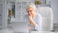 Tired sleepy old lady manager types on grey laptop at white table sitting on large chair against different shelves