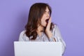 Tired sleepy woman yawning and covering mouth with hand, working on laptop sitting against lilac background, wearing elegant