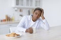 Tired sleepy young attractive black woman with cup of drink sits at table with cookies in scandinavian kitchen interior Royalty Free Stock Photo