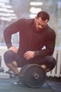 Tired strong young caucasian man with beard in black sportswear sit with iron barbell relaxing after heavy intensive workout