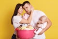 Tired troublesome parents crying newborn baby and basin full of clean clothing, have much domestic work, posing isolated on yellow