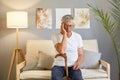 Tired unhealthy grey-haired senior man in casual white T-shirt sitting on couch at home in living room interior suffering headache