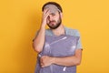 Tired unshaven man standing against yellow background, holding grey pillow, touching his head, wearing gray t shirt and blindfold