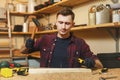 Handsome smiling young man working in carpentry workshop at wooden table place with piece of wood Royalty Free Stock Photo