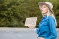 Tired woman hopefully look out passing cars with cardboard poster on roadside in forest. Lady in hat and denim outfit