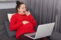 Tired woman keeps hand on belly, anticipates for baby, yawns as watches film online, focused into laptop computer, leans on