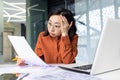 Tired young chinese woman wearing glasses sitting at desk in office with pile of papers in hands, holding head from Royalty Free Stock Photo