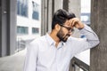 Tired young Indian man standing near a building, leaning against a wall and holding his head with his hand, suffering Royalty Free Stock Photo
