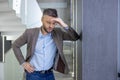 Tired young man standing by the wall in the office and holding his head, feeling bad, worried Royalty Free Stock Photo