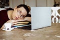 Tired young woman in glasses lying down on books and looking at laptop screen at desk in home room. Tiredness from