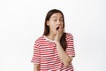 Tired young woman yawning, feeling exhausted or sleepy, waking up early in morning, standing in red t-shirt against