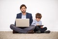 Today is online bonding time. A father and son sitting on the floor against a wall with a laptop and digital tablet. Royalty Free Stock Photo