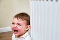 Toddler baby crying at the radiator, tearful child face close-up. White ra