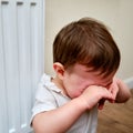 Toddler baby crying at the radiator, tearful child face close-up. White ra