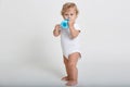 Toddler boy drinking water from blue feeding bottle, child wearing white bodysuit, looking away, standing barefoot against light Royalty Free Stock Photo