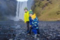 Toddler boy and siblings, playing with rocks on the river in fronf of Skogafoss waterfall in Iceland on a sunset cloudy day