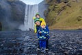 Toddler boy and siblings, playing with rocks on the river in fronf of Skogafoss waterfall in Iceland on a sunset cloudy day