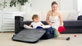 Cute toddler boy sitting in suitcase while mother packing things in suitcase for holiday