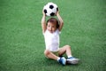 Toddler boy in sports uniform sitting on green footall field holding soccer ball above head. Copy space Royalty Free Stock Photo