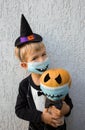 Toddler boy in witch hat holds pumpkin with black eyes . Both wear protective masks with horrible smile painted on them Royalty Free Stock Photo