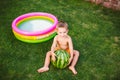 Toddler cute Caucasian white European race boy sitting hugging holding huge watermelon and smiling on background of green grass,