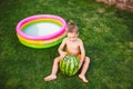 Toddler cute Caucasian white European race boy sitting hugging holding huge watermelon and smiling on background of green grass,