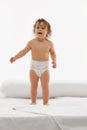 Toddler, little girl in diaper standing on white bed, looking curious and playful against white background. Smiling Royalty Free Stock Photo
