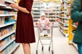 A toddler sits in a grocery cart and looks around while his mother picks out groceries. Family shopping in the supermarket