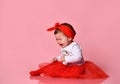 Toddler in white bodysuit, red headband, poofy skirt. She crying, sitting on floor against pink studio background. Close up