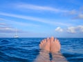 Feet floating on surface of sea with boat on horizon under blue