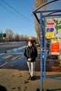 A red-haired girl in a medical mask stands at a public transport stop on a sunny spring evening.