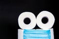 Toilet paper rolls with a medical mask folded in the form of an emoticon, on a black background. concept of panic during