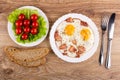 Tomato on lettuce in plate, fried eggs with pork brisket in plate, pieces of bread, fork and knife on table. Top view Royalty Free Stock Photo